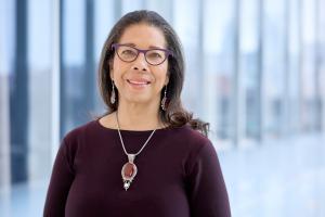 Alexis A. Thompson smiling, wearing a deep purple colored sweater, purple glasses, and matching earrings and necklace set.