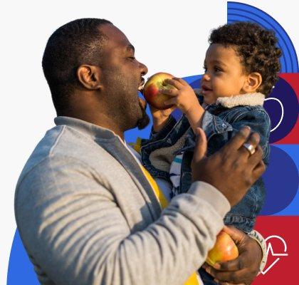 A dad holds his young child while the child holds an apple up to his mouth for him to bite into.