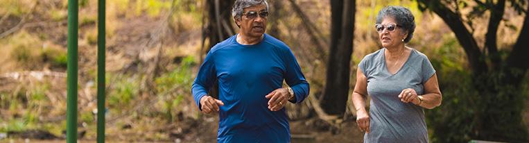 An older couple jogs on a path through a wooded park.