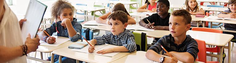 Children sitting at desks listen to their teacher while writing in notebooks with pencils.