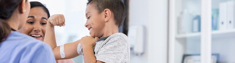 A young boy with a bandage on his arm flexes his muscles for a health care provider and his mom.