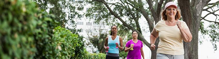 Three women, two of them holding small weights, walk briskly in a park.